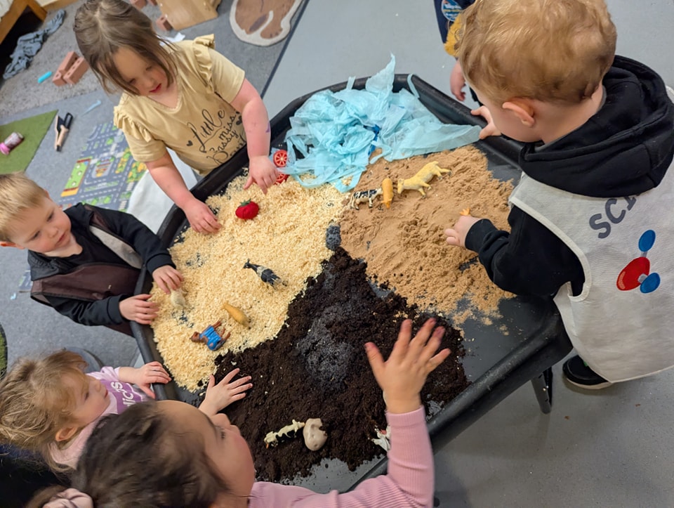 Group of young children standing around a table and playing with different coloured sand